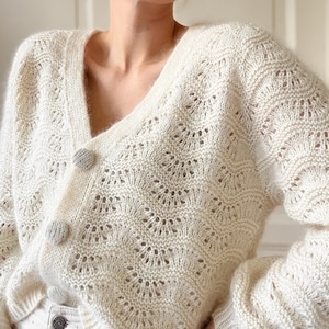 May include: A white crocheted cardigan with a V-neck and button closure. The cardigan has a delicate openwork pattern.