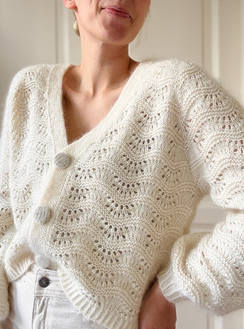 May include: A white crocheted cardigan with a V-neck and button closure. The cardigan has a delicate openwork pattern.