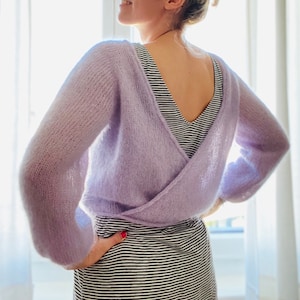 Könnte beinhalten: Ein lila gestrickter Schal mit einem Kreuz-Design auf der Vorderseite. Der Schal wird über einem schwarz-weiß gestreiften Shirt getragen.