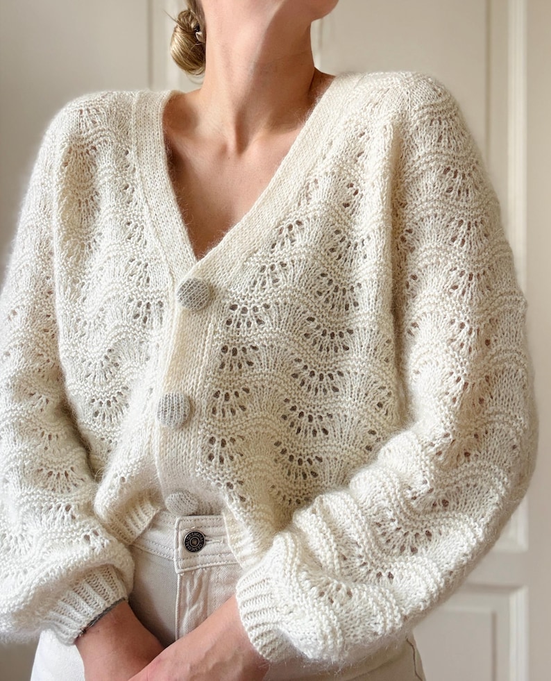 May include: A white crocheted cardigan with a V-neck and button closure. The cardigan has a delicate openwork pattern and long sleeves.