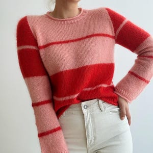 Könnte beinhalten: Ein Rundhals-Pullover mit horizontalen Streifen in Rot- und Rosatönen. Der langärmlige Pullover ist aus einem weichen, flauschigen Material gefertigt. Das Model trägt eine helle Jeans.