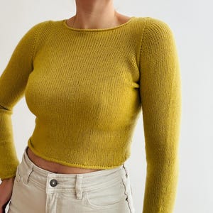 May include: Mustard yellow long-sleeved sweater with a crew neck. The cropped sweater is paired with light beige jeans. The sweater is made of a knitted material and has a ribbed texture.