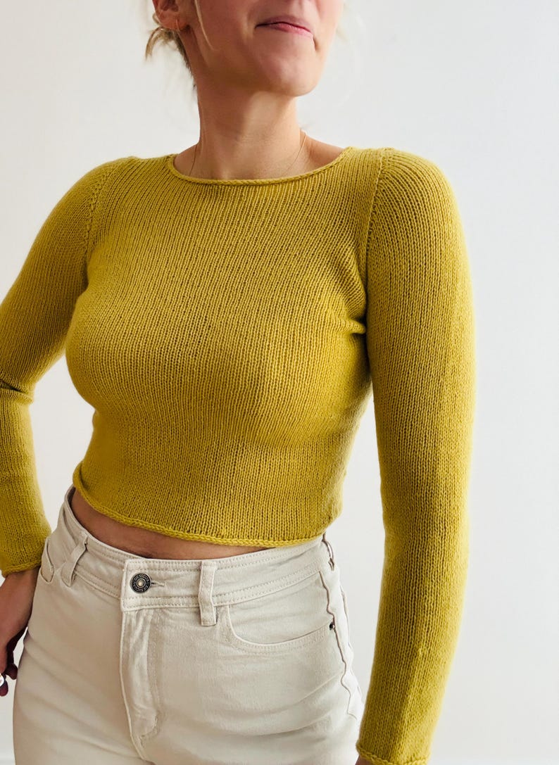 May include: Mustard yellow long-sleeved sweater with a crew neck. The cropped sweater is paired with light beige jeans. The sweater is made of a knitted material and has a ribbed texture.