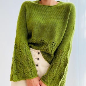 May include: A green knitted jumper with bell sleeves. The jumper has a round neckline and a decorative openwork pattern on the lower sleeves. The person is wearing a cream-coloured skirt with buttons.