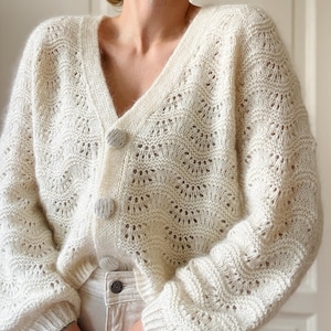 May include: A white crocheted cardigan with a V-neck and button closure. The cardigan has a delicate openwork pattern and long sleeves.