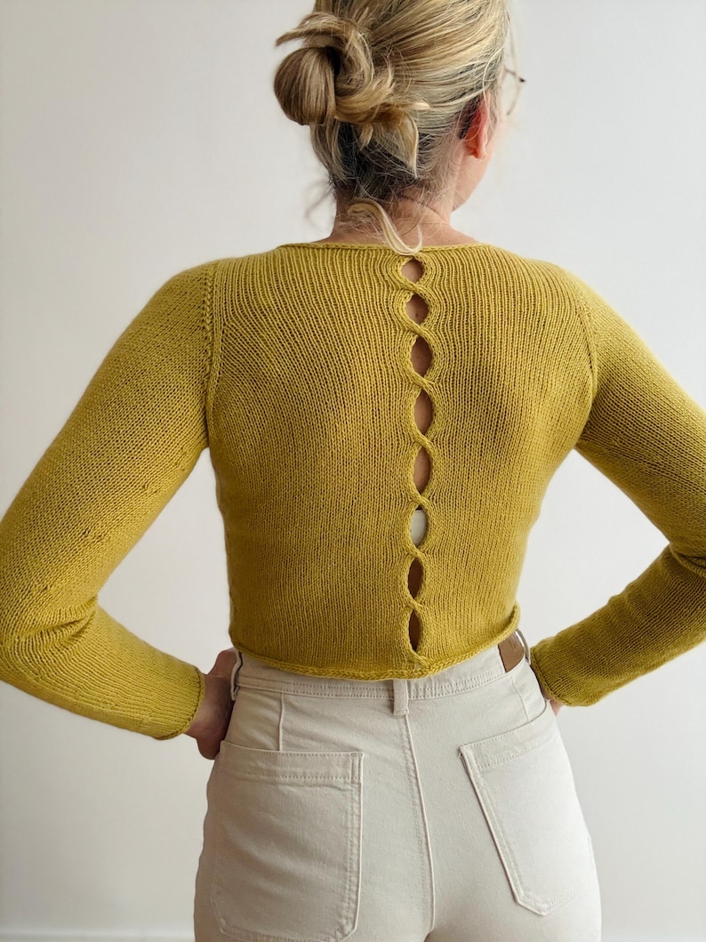 May include: Mustard yellow long-sleeved sweater with a unique open back design. The back features a series of vertical, lozenge-shaped cutouts. The sweater has a crew neck and is made of a knitted material. The model is wearing light-colored pants.