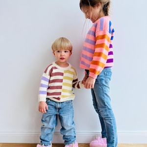 Può includere: Due bambini con maglioni a righe colorati e jeans blu. Il più piccolo indossa un maglione bicolore e entrambi indossano pantofole a forma di unicorno rosa. I maglioni presentano strisce orizzontali.