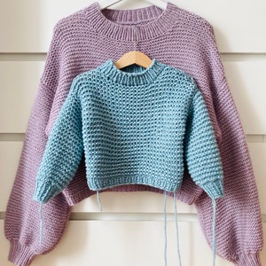 Può includere: Due maglioni di lana appesi a un appendiabiti bianco. Il maglione superiore è di colore lilla chiaro e il maglione inferiore è di colore azzurro chiaro.