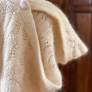 May include: A cream-colored knitted shawl with a delicate lace pattern and a single button closure.