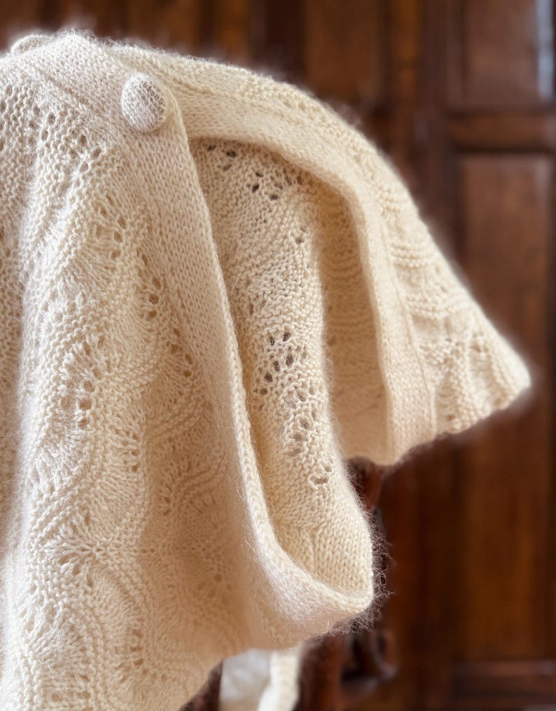 May include: A cream-colored knitted shawl with a delicate lace pattern and a single button closure.