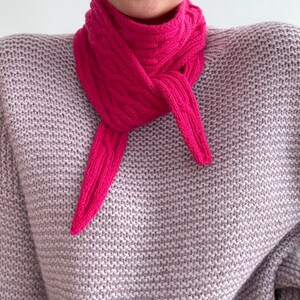Könnte beinhalten: Ein leuchtend pinkfarbener Strickschal ist um den Hals gebunden, die Enden hängen herunter. Der Schal ist aus einem dicken, strukturierten Garn gefertigt. Die Person trägt einen hellgrauen Pullover. Der Schal setzt einen Farbakzent.