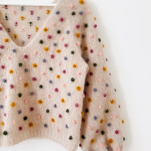 May include: A beige knitted sweater with a V-neck and long sleeves. The sweater is covered in small, colorful pom-poms.