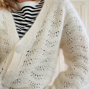 May include: A white crocheted cardigan with a delicate openwork pattern. The cardigan has a V-neck and long sleeves.