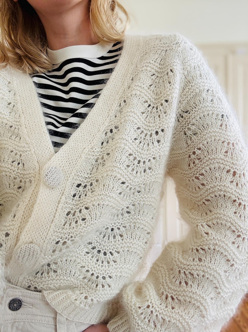 May include: A white crocheted cardigan with a delicate openwork pattern. The cardigan has a V-neck and long sleeves.