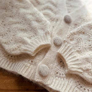 May include: A white knitted cardigan with a delicate openwork pattern and three beige buttons.