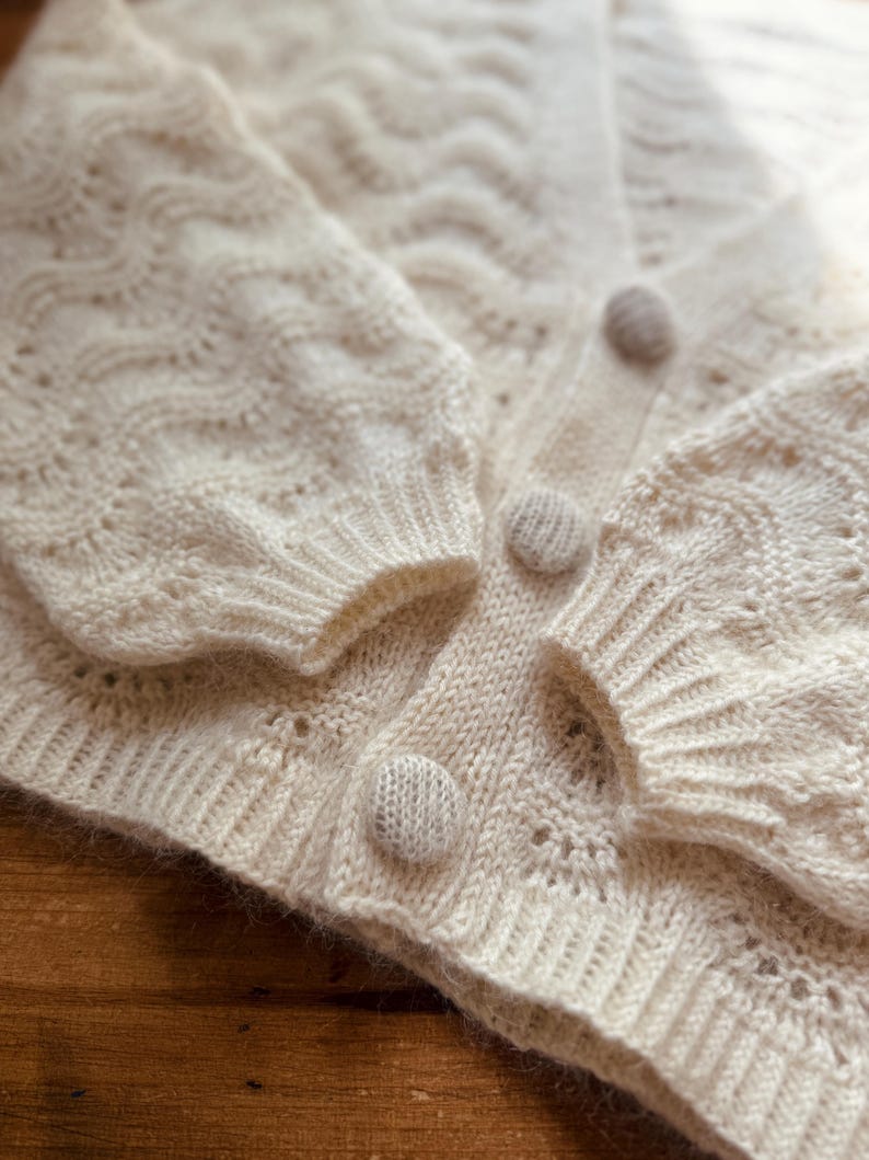 May include: A white knitted cardigan with a delicate openwork pattern and three beige buttons.