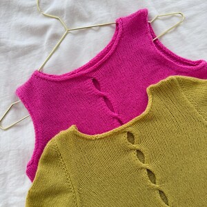 May include: Two knitted tops in vibrant colors. One is a bright pink sleeveless top hanging on a gold hanger. The other is a long-sleeved olive green top with a decorative opening down the front. Both are made of a soft, textured knit.
