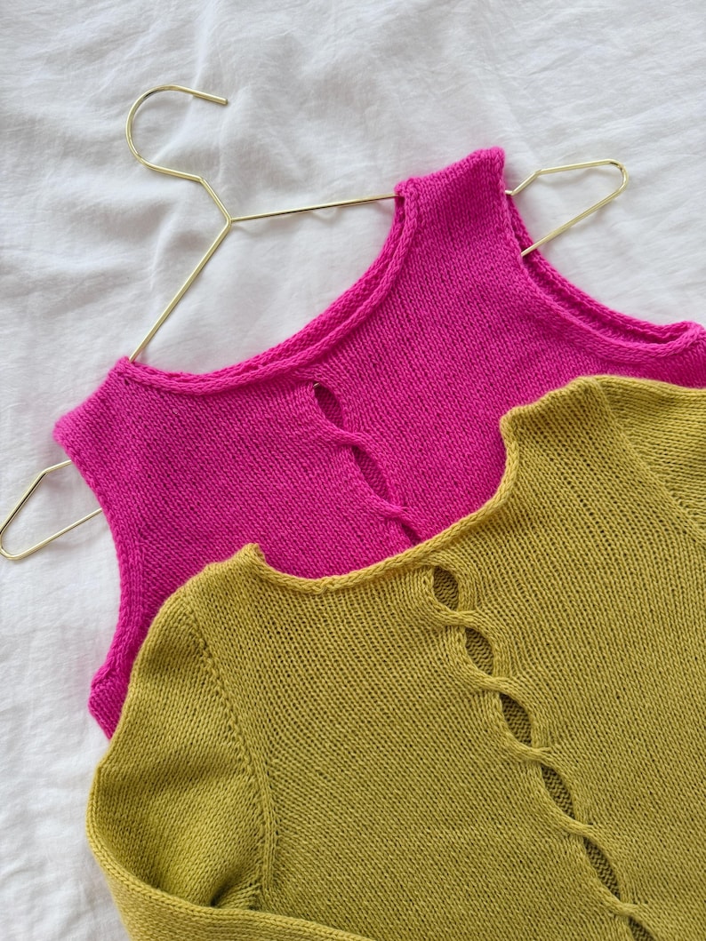 May include: Two knitted tops in vibrant colors. One is a bright pink sleeveless top hanging on a gold hanger. The other is a long-sleeved olive green top with a decorative opening down the front. Both are made of a soft, textured knit.