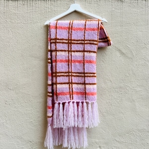 Könnte beinhalten: Ein rosa-brauner karierter Schal mit Fransen-Quasten, der an einem weißen Kleiderbügel an einer hellgrauen Wand hängt.