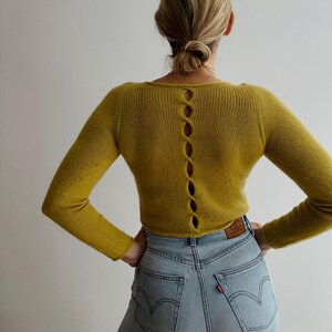 May include: Mustard yellow long-sleeve sweater with a scoop neck and a series of oval-shaped cutouts down the back. The sweater is paired with light wash blue jeans.
