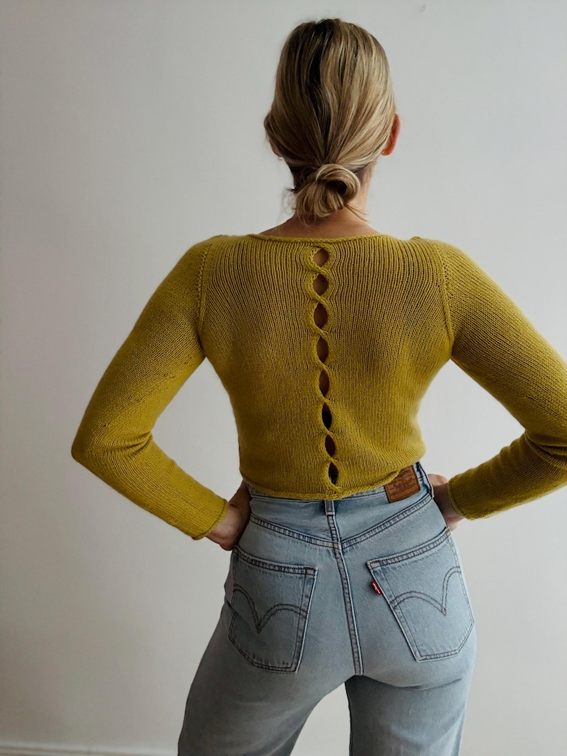 May include: Mustard yellow long-sleeve sweater with a scoop neck and a series of oval-shaped cutouts down the back. The sweater is paired with light wash blue jeans.