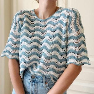 Peut inclure: Un haut en tricot à manches courtes et à tissage ouvert, avec un motif de vagues bleu clair et crème. Le haut a une encolure dégagée et est associé à un jean en denim délavé. Le motif en tricot crée un look délicat et aéré.