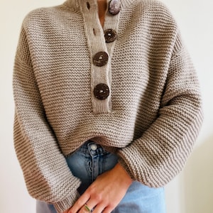 Könnte beinhalten: Ein beiger Strickpullover mit Stehkragen und einer Reihe brauner Holzknöpfe auf der Vorderseite.