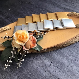May include: A wooden tray with a natural bark edge, decorated with a cluster of artificial flowers and a row of gold and silver wrapped chocolates.