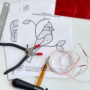 May include: A stained glass suncatcher pattern of a red cardinal bird with numbered sections for glass pieces. The pattern is printed on white paper and includes a pair of pliers, a glass cutter, copper foil tape, and a sheet of red glass.