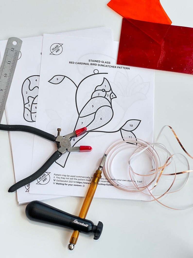 May include: A stained glass suncatcher pattern of a red cardinal bird with numbered sections for glass pieces. The pattern is printed on white paper and includes a pair of pliers, a glass cutter, copper foil tape, and a sheet of red glass.
