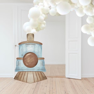 Puede incluir: Un tren de juguete azul y marrón con una nube de globos blancos encima. El tren está en un suelo de madera delante de una puerta blanca.