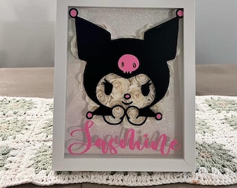 Puede incluir: Una obra de arte enmarcada con un personaje de dibujos animados negro con una calavera y acentos rosas. El personaje está sobre un fondo de flores de papel blanco y purpurina. El nombre "Sunshine" está escrito en rosa en la parte inferior.