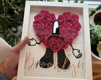 Puede incluir: Un marco blanco con un corazón de flores de papel rosa, brazos y piernas de dibujos animados negros y el nombre "Brooklyn" escrito en purpurina dorada.