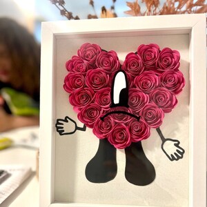Puede incluir: Un marco blanco con un coraz&oacute;n de flores de papel rosa con una silueta negra de una persona con una cara triste. La persona tiene brazos y piernas y lleva un atuendo negro.