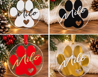 Puede incluir: Cuatro adornos circulares de acrílico transparente con un diseño de huella de pata y un recorte en forma de corazón. Cada adorno muestra el nombre Milo en letra cursiva. Las huellas están disponibles en blanco, negro, rojo y dorado.