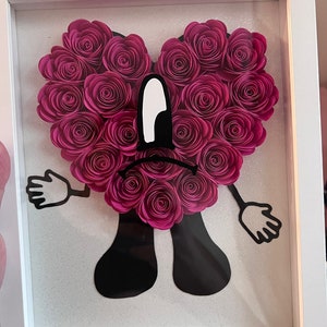 Puede incluir: Una obra de arte enmarcada que presenta un coraz&oacute;n de flores de papel rosa con una cara de dibujos animados negra. La cara tiene un ojo blanco grande y una mueca.