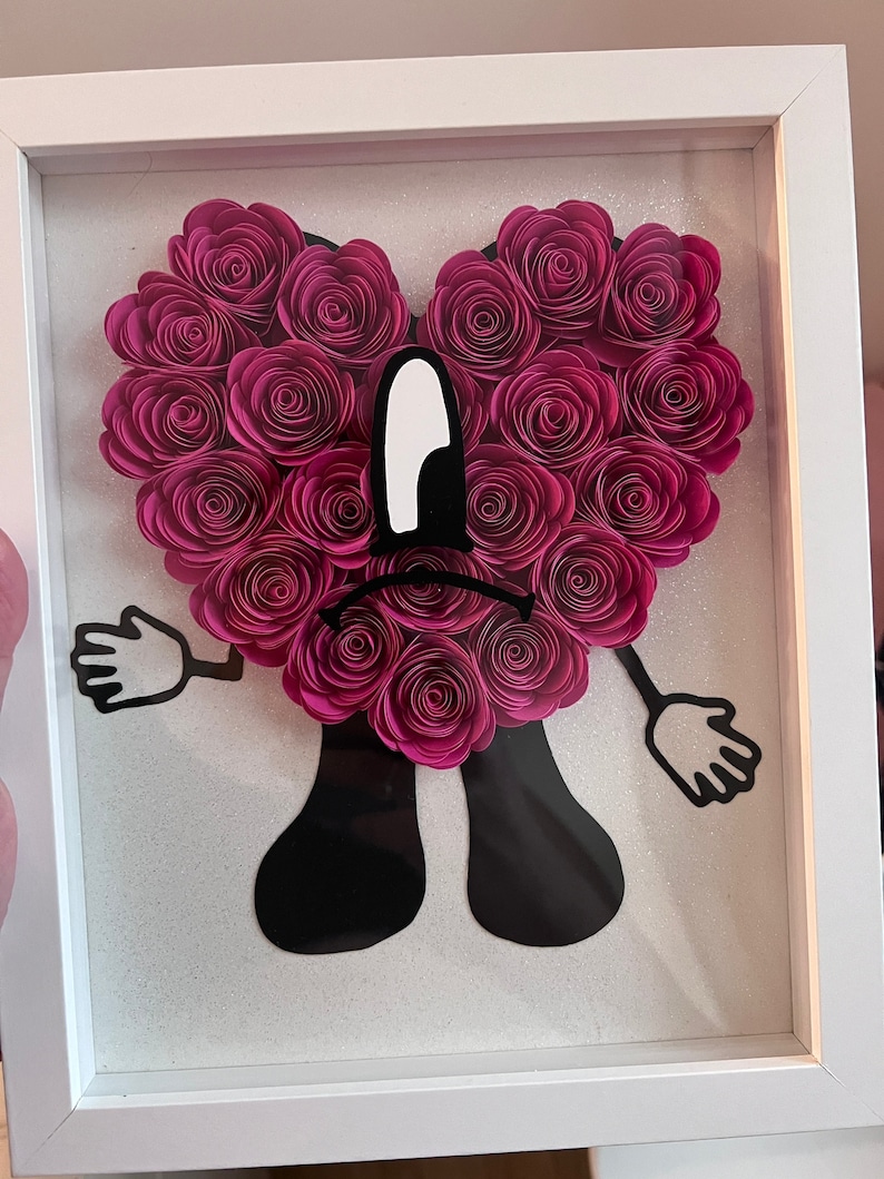 Puede incluir: Una obra de arte enmarcada que presenta un coraz&oacute;n de flores de papel rosa con una cara de dibujos animados negra. La cara tiene un ojo blanco grande y una mueca.