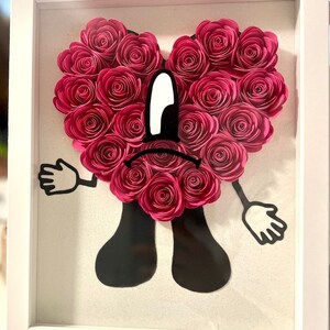Puede incluir: Una obra de arte de papel enmarcada que presenta un personaje de coraz&oacute;n rosa con una expresi&oacute;n triste. El coraz&oacute;n est&aacute; hecho de rosas de papel rosa y el personaje tiene brazos y piernas negros.