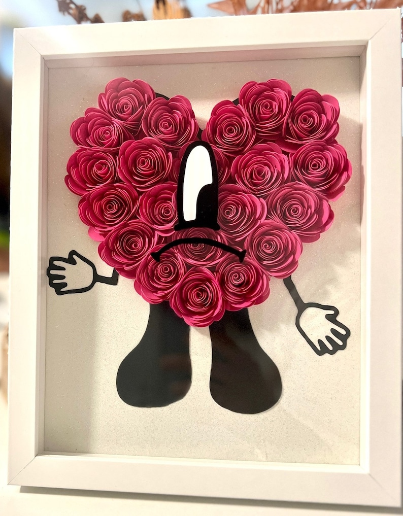 Puede incluir: Una obra de arte de papel enmarcada que presenta un personaje de coraz&oacute;n rosa con una expresi&oacute;n triste. El coraz&oacute;n est&aacute; hecho de rosas de papel rosa y el personaje tiene brazos y piernas negros.