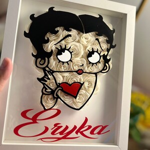 Puede incluir: Un marco blanco con una imagen en blanco y negro de un personaje de dibujos animados con rosas de papel blanco y un coraz&oacute;n rojo. El nombre "Eryka" est&aacute; escrito en rojo cursivo debajo de la imagen.
