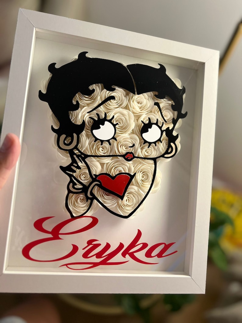Puede incluir: Un marco blanco con una imagen en blanco y negro de un personaje de dibujos animados con rosas de papel blanco y un coraz&oacute;n rojo. El nombre "Eryka" est&aacute; escrito en rojo cursivo debajo de la imagen.