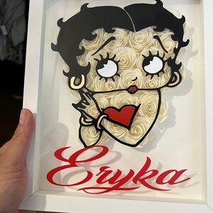 Puede incluir: Un marco blanco con una imagen en blanco y negro de un personaje de dibujos animados con un coraz&oacute;n rojo. El nombre del personaje, "Eryka", est&aacute; escrito en letras rojas cursivas debajo de la imagen.
