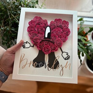 Puede incluir: Un marco blanco con un coraz&oacute;n de flores de papel rosa, brazos y piernas negros, y el nombre "Brooklyn" escrito en purpurina dorada.