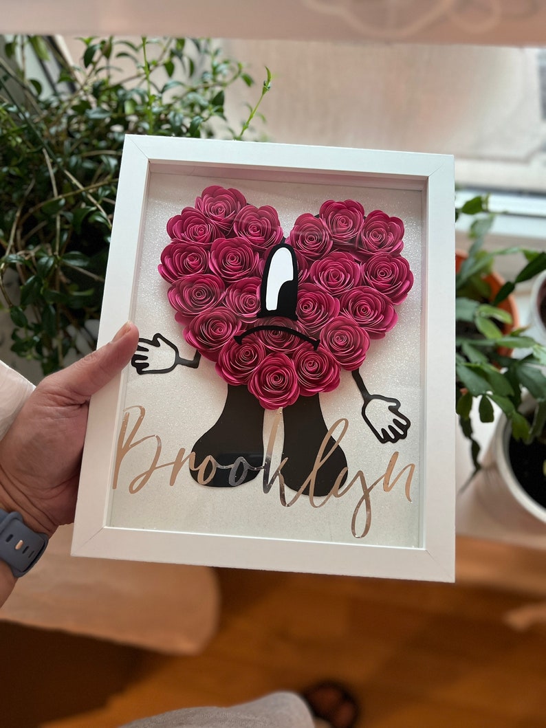 Puede incluir: Un marco blanco con un coraz&oacute;n de flores de papel rosa, brazos y piernas negros, y el nombre "Brooklyn" escrito en purpurina dorada.