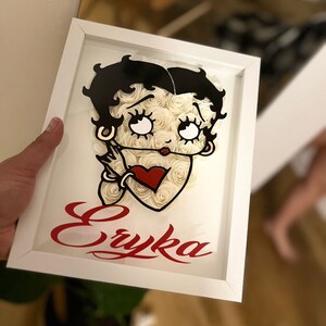 Puede incluir: Un marco blanco con una imagen en blanco y negro de Betty Boop hecha de rosas de papel blanco. El nombre "Eryka" est&aacute; escrito en letras rojas cursivas debajo de la imagen.