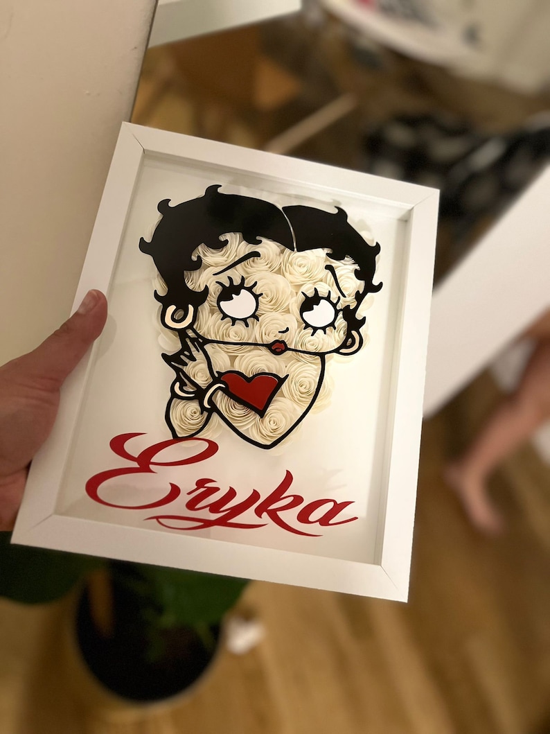 Puede incluir: Un marco blanco con una imagen en blanco y negro de Betty Boop hecha de rosas de papel blanco. El nombre "Eryka" est&aacute; escrito en letras rojas cursivas debajo de la imagen.