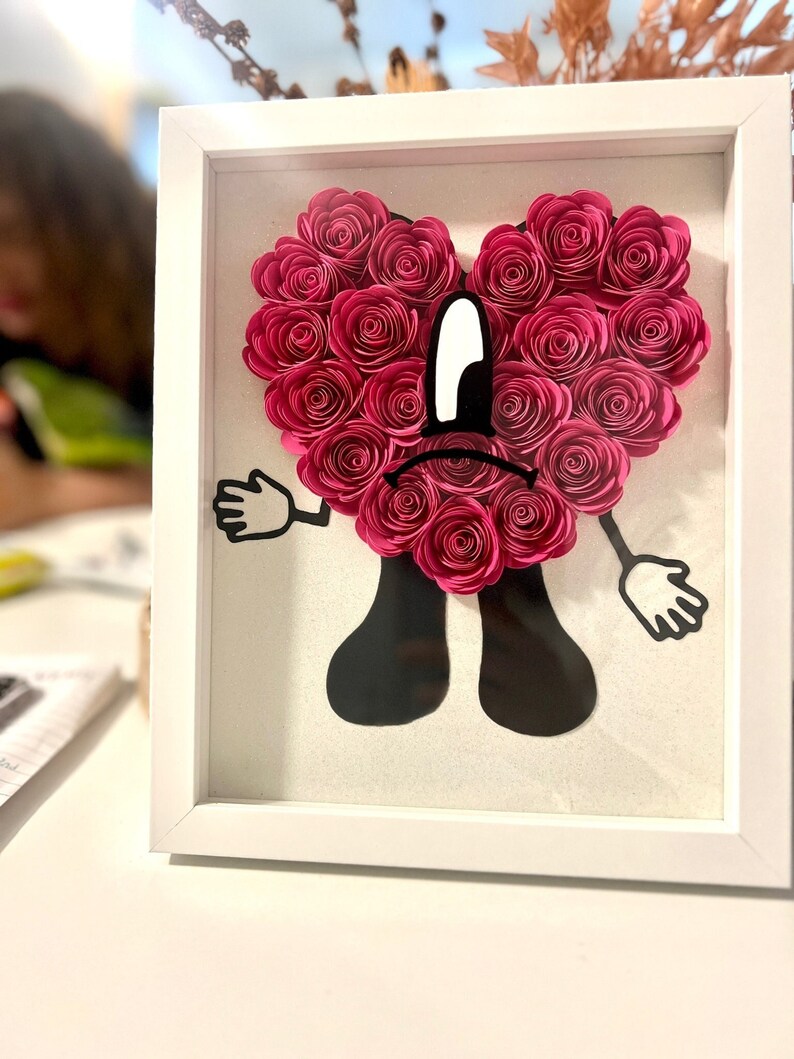 Puede incluir: Un marco blanco con un coraz&oacute;n de flores de papel rosa con una silueta negra de una persona con una cara triste. La persona tiene brazos y piernas y lleva un atuendo negro.