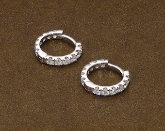 May include: Two small, silver hoop earrings with a row of clear, sparkling stones. The earrings are displayed on a brown surface, showcasing their delicate design and reflective qualities. These earrings are a classic jewelry item.