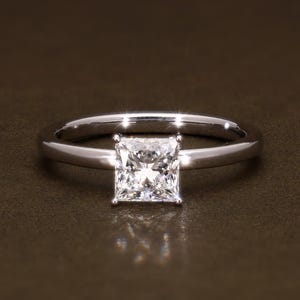 Könnte beinhalten: Ein silberner Ring mit einem quadratisch geschliffenen Diamanten als Mittelstein. Der Ring hat ein schlichtes Band, und der Diamant funkelt hell. Der Ring wird auf einer braunen Oberfläche präsentiert.