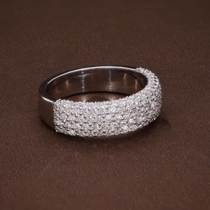 May include: A silver ring with a band encrusted with small, clear gemstones. The ring is set against a brown background, showcasing its sparkling details. The gemstones cover the top and sides of the ring.